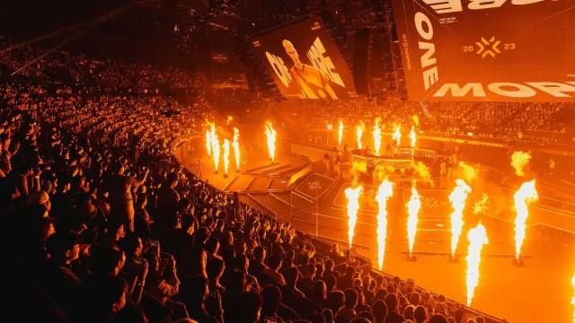 Een enorme menigte kijkt naar grote schermen met de tekst "ONE MORE".  Hun silhouetten worden verlicht door meerdere vuurkolommen.